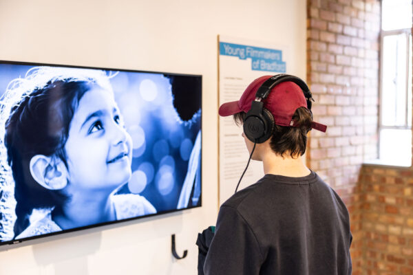 A person wearing a cap and a pair of headphones, watches a film on a screen.