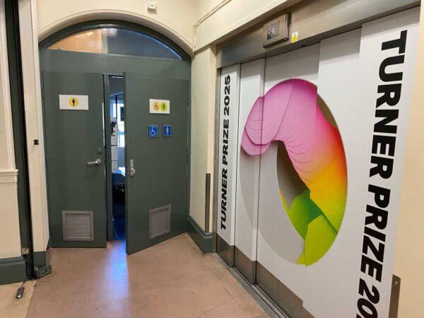 On the left there is a grey door with an accessible toilet sign on it, showing a wheelchair in a circle. There is also a baby changing sign. On the right there is a lift, with Turner Prize branding on the doors.