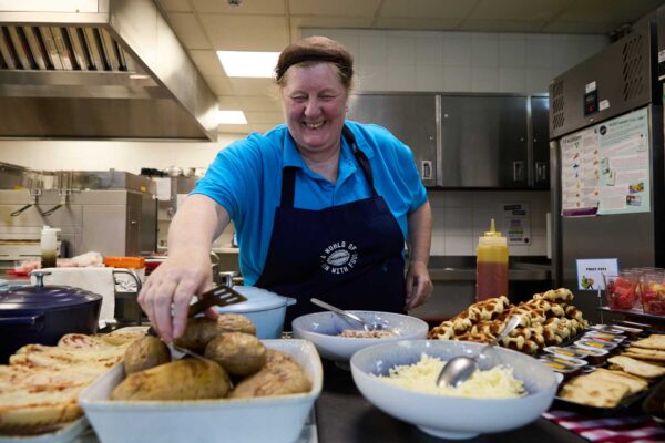 A smiling woman in a blue apron and hairnet. She is working in a kitchen.