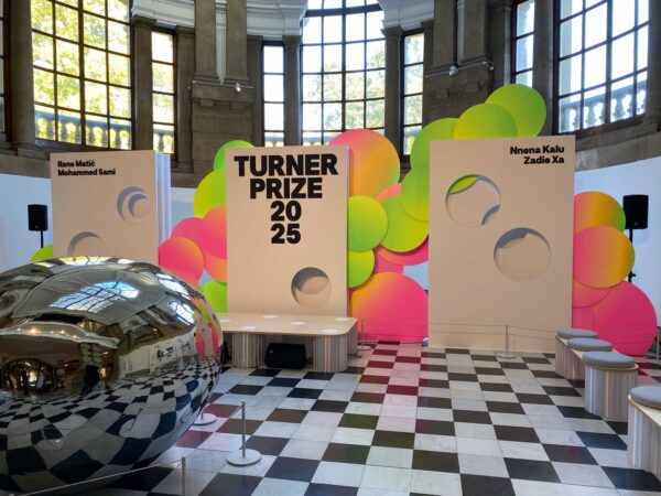 A room with black and white checkerboard flooring and three large signs - the signs read Turner Prize 2025 and have the four names of the Turner Prize artists on them. Behind the signs are large colourful circles. In the foreground a large, round, reflective artwork is on the floor.