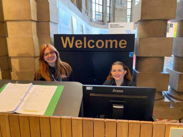 Two smiling people behind a computer screen. There is a sign behind them that says Welcome.