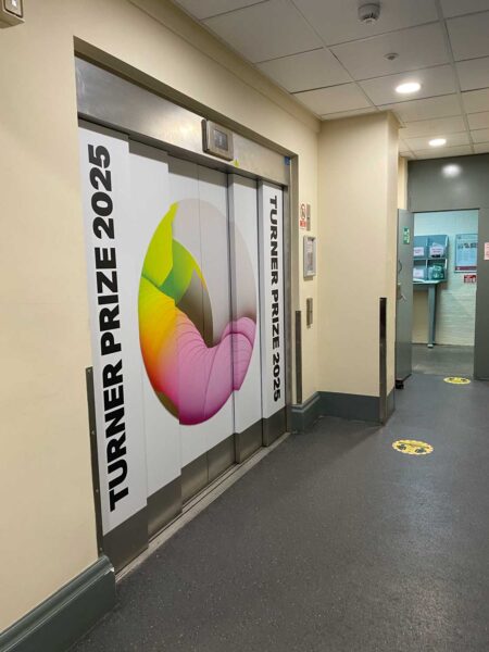 A lift on the left side of the image. This has branding on it which reads Turner Prize 2025 and has some bright pink and green colours on it.
