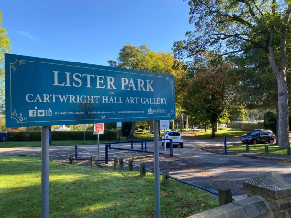 A blue metal sign with white writing. The writing reads Lister Park and Cartwright Hall Art Gallery. There are some autumn trees behind the sign and grass underneath it.