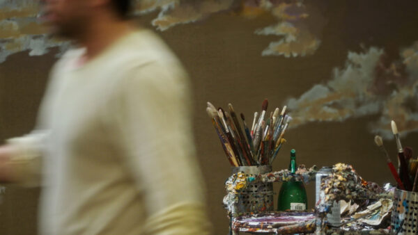 An image of artist Mohammed Sami walking past us - he appears blurred, and we only see his torso in a white top. In focus behind him is a table with paint brushes and dried paint.