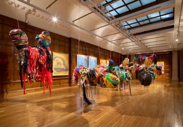 A view inside Nnena Kalu's Turner Prize installation. There are large hanging sculptures around the room made of tape, in red, green, yellow, black and more colours. Along the walls we see hanging drawings.
