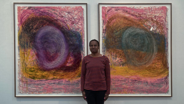 Artist Nnena Kalu stands with her arms by her side in front of two paintings. These abstracit paintings have swirled shapes created in repetitive patterns, in pink, purple, and orange colours.