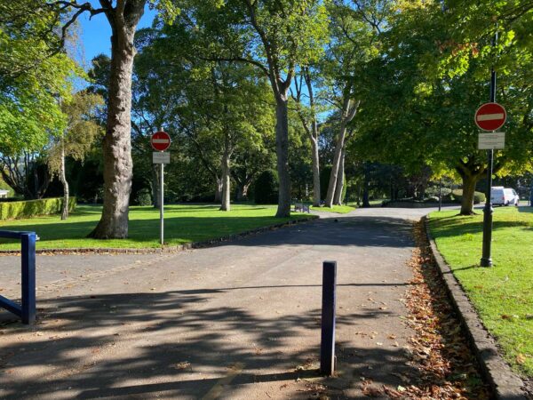 A small road lined with trees. There is a blue bollard to the right, and just at the left of the picture there is an open barrier. In front of us are two red and white no entry signs.