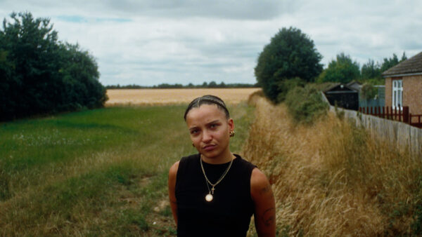 Artist Rene Matic stands in a field, close to a brick house. They look up towards us wearing a black top with gold chains around their neck.