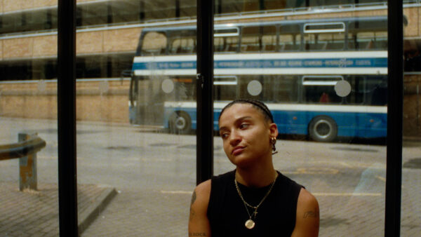 Artist Rene Matic sits in an urban space, in a bus stop. They look quietly out with a solemn expresion.