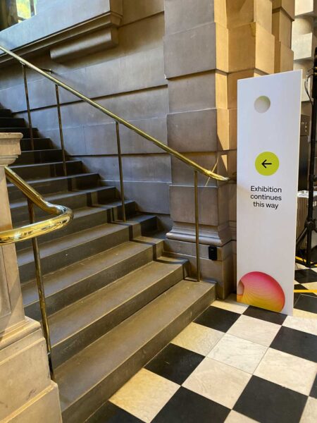 A set of stone stairs, with a sign next to them that reads Exhibition continues this way, with an arrow pointing up the stairs.