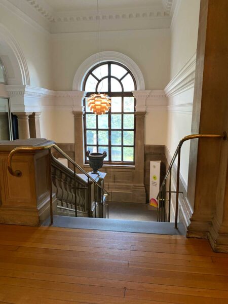 A stone staircase leading down, with a brass handrail. There is a large window in front of us.