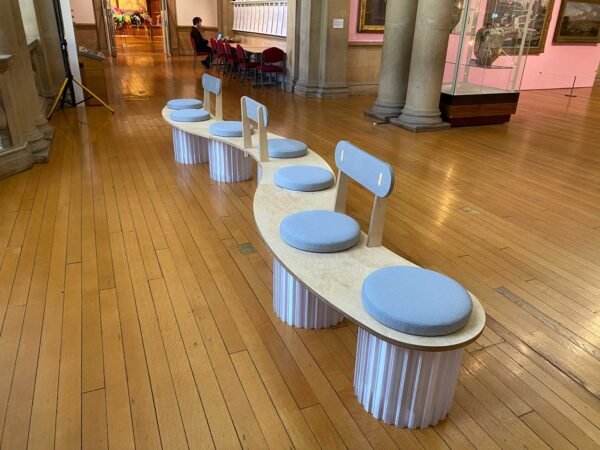A bench made of wood - it is a wavy piece of wood with several round blue cushions on it and three back rests.