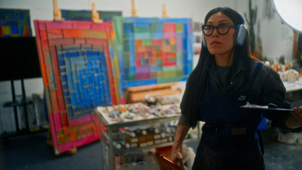 Artist Zadie Xa wears headphones as seh works in studio. She holds a palette and paint brush.