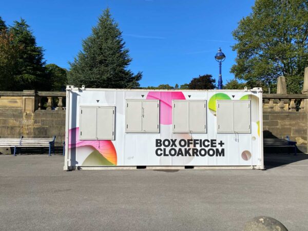 A shipping container with four windows. One of the windows is lower than the others. There is text on the shipping container that reads in all capitals BOX OFFICE + CLOAK ROOM. The box is mostly white but there are pink and green colours too - the Bradford 2025 and Turner Prize brand colours.