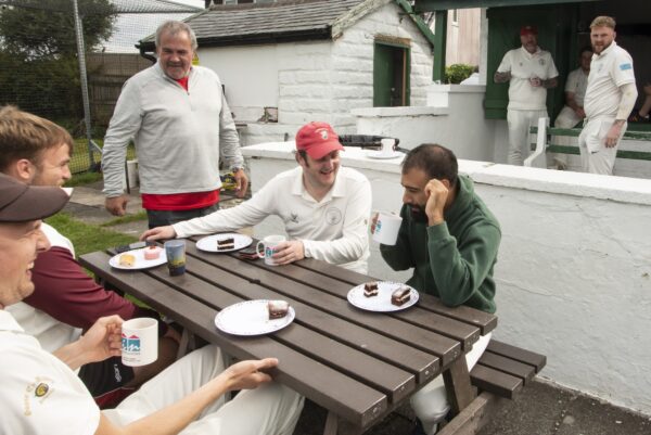 Men sat outside of the cricket club enjoying eachothers company and food.