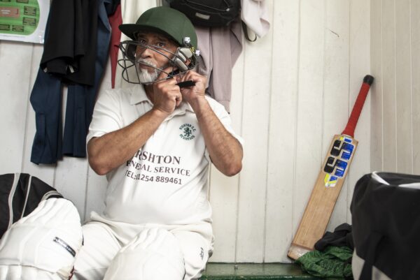 Man in cricket gear, sat in the changing rooms putting on his head gaurd. There is a bat sat next to him.