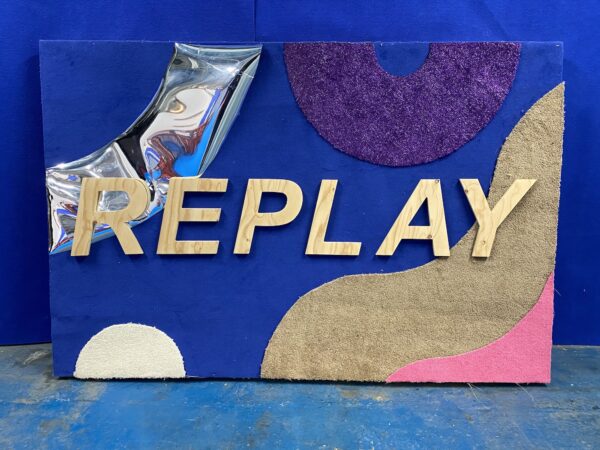 A collage-style REPLAY sign made out of fabric and wood.
