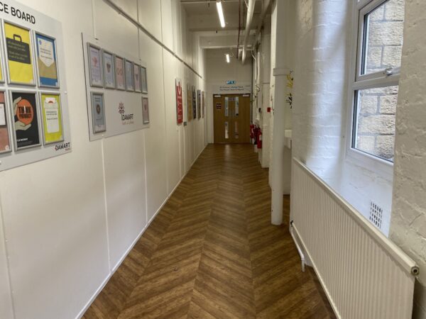A corridor with a wooden floor and cream walls with a window and notice boards on it.