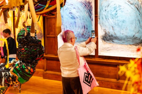 A person with pink hair takes a photograph inside an art exhibition.