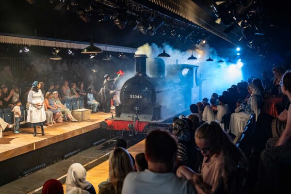 A steam train enters the auditorium at The Railway Children, with members of the audience sitting either side of the tracks.