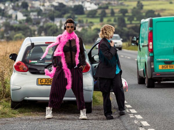 A portrait photo of two best mates and performance/live art makers, Jess Murrain and Lua Bairstow. There is a small silver car with a Palestine sticker on its window, parked beside a road on the edge of the Baildon Moor in Bradford, West Yorkshire. Jess, a brown person with blonde and brown afro hair, is standing in front of the back of the car. Posed, looking directly at the camera. Wearing white Doc Martens, grey trousers with pink pinstripes, a sheer black shirt, a vivid pink feather boa, sunglasses and headphones. The door of the car is open. Lua, a white, gender non-conforming person, is standing next to Jess and beside the open car door, looking back at the road behind them. Wearing a pinstriped suit with a turquoise shirt just peeking through.