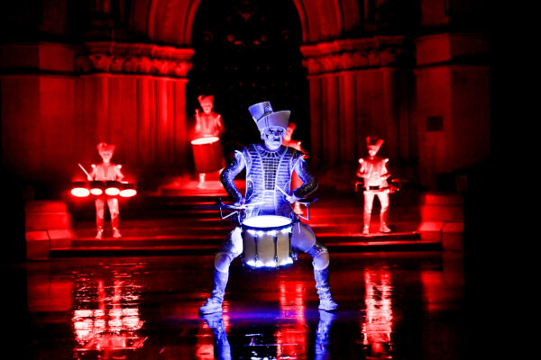 Men light up in red and blue coloured lighting on a dark night, dressed in all white doing a drumming performance