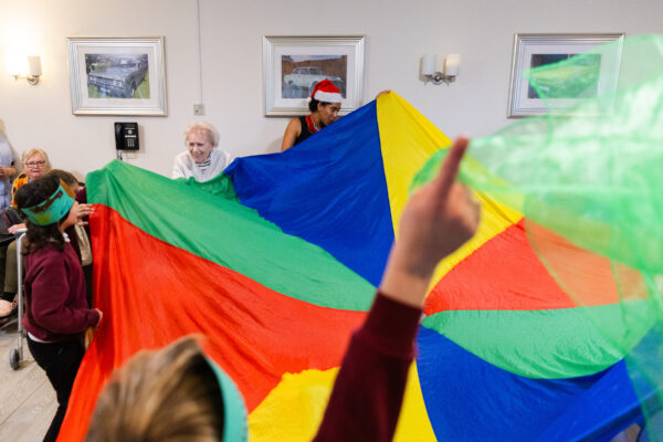 Young children and elderly people all playing with a rainbow colour parachute and colourful balls in a care home