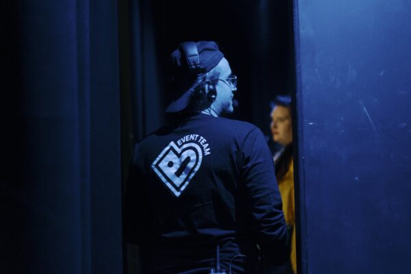 A man behind stage wiht a headset on and wearing an event t-shirt