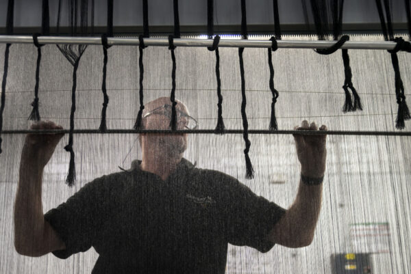 A man weaving. He stands behind the woollen yarn holding a stick horizontally.
