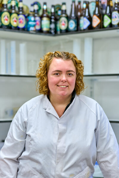 A woman with blonde hair and a white lab coat sits in a lab. Beer bottles on a high shelf are behind her.