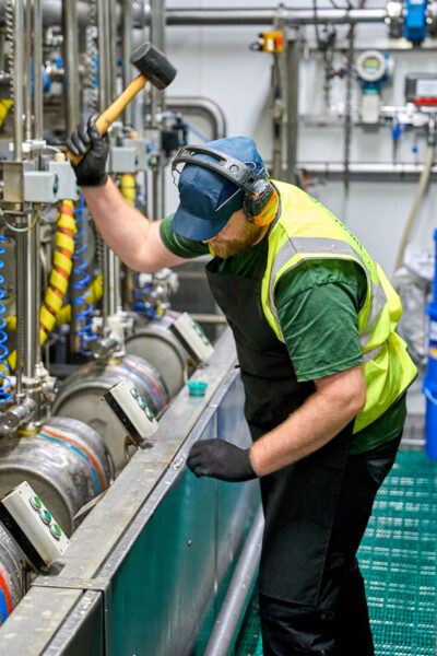 A man with orange headphones and a blue cap using a hammer in a factory.