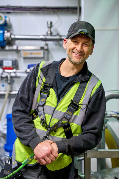 A man in a cap and harness smiles in a factory.