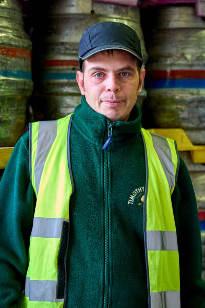 A man with blue eyes in a hives and green fleece, with beer caskets in the background.
