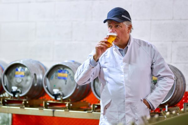 An older man in a white jacket and blue cap, drinks a half pint of beer.