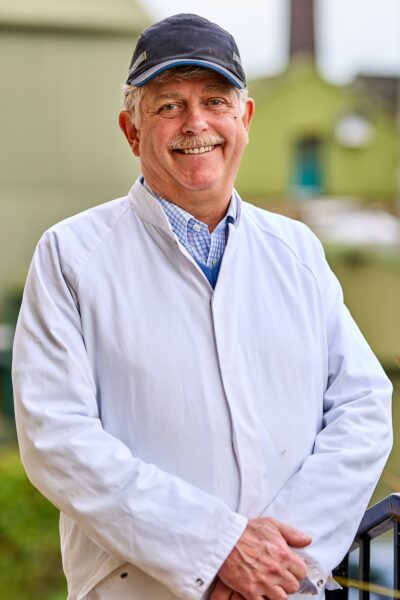 A man smiling in a white lab coat and blue cap posing to the camera.