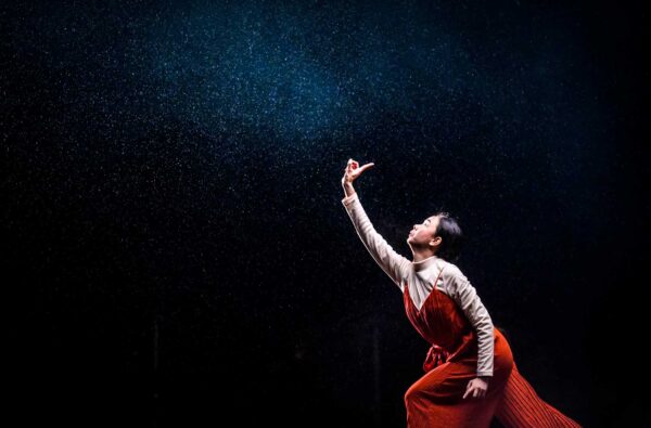 A lone performer in a red and white costume. She holds her finger high in the air against a black background.
