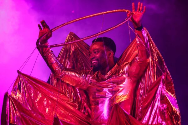 A smiling performer raises his hands in the air. He wears a shiny costume with a large ring above his head.