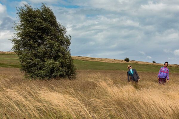 A photo of two best mates and performance/live art makers, Jess Murrain and Lua Bairstow. They are in a field. There is a tree. Jess, Lua and the tree are leaning with and against the breeze.