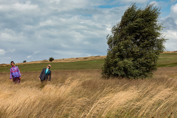 A photo of two best mates and performance/live art makers, Jess Murrain and Lua Bairstow. They are in a field. There is a tree. Jess, Lua and the tree are leaning with and against the breeze.