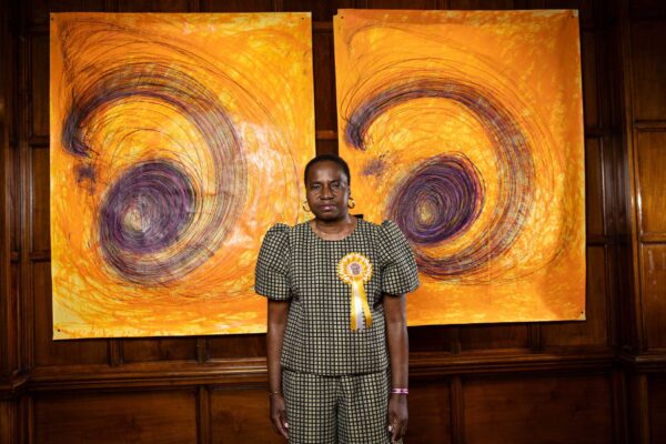 Artist Nnena Kalu stands in front of her artwork - two drawings with spirals on an orange yellow background.