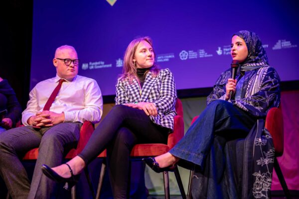 Three young people on a stage. They are sitting. One of them holds a microphone.