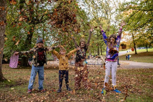 Four children stand in a line in a woodland, throwing leaves into the air.