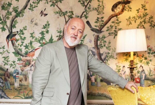 Comedian Bill Bailey wears a pale suit, standing in an opulent room. He leans on a chair.