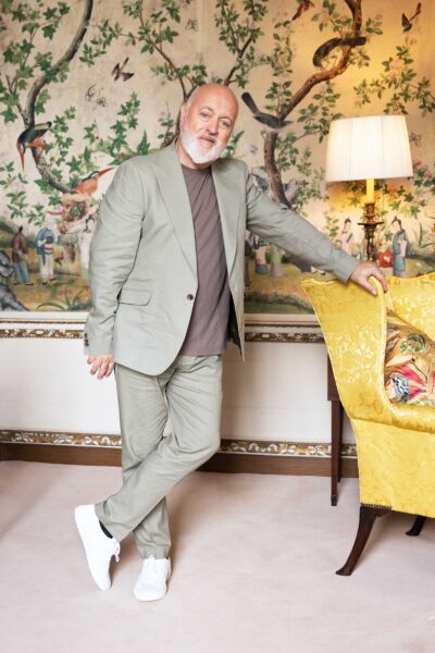 Comedian Bill Bailey wears a pale suit, standing in an opulent room. He leans on a chair.